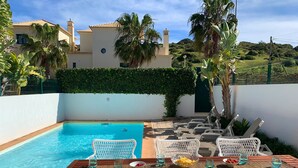 A poolside area with a wooden table, white wicker chairs, and a view of a house with a tiled roof and a green hedge.