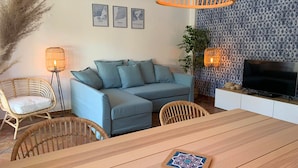A modern living room with a sofa, two wicker chairs, a wooden dining table, a TV, and decorative wall art.