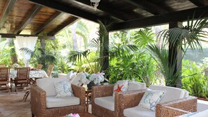 A covered outdoor seating area with wicker furniture, a wooden table, and potted plants.