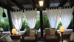 A covered outdoor seating area with wicker furniture and white cushions, a wooden coffee table with a floral arrangement, and a stone wall in the background.