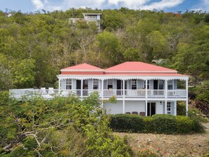 A two-story white building with a red roof, surrounded by dense greenery and trees.