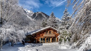 A wooden cabin with a balcony, surrounded by snow-covered trees and mountains in the background.
