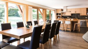 A modern kitchen with wooden cabinets, a dining area with a wooden table and chairs, and large windows offering a view of the outdoors.