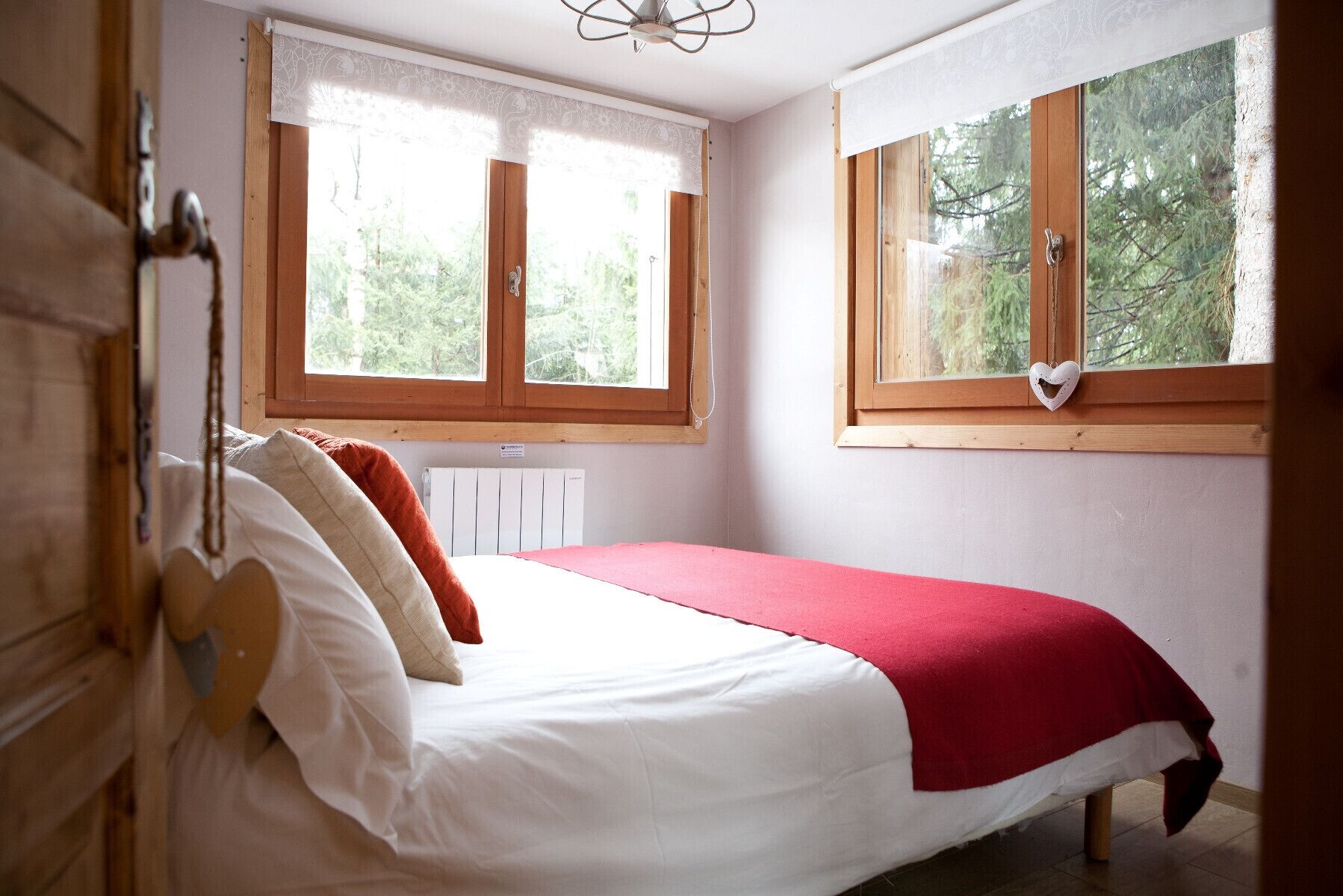 Een slaapkamer met een bed, twee ramen met gordijnen, een radiator en een hartvormige decoratie op een raam.