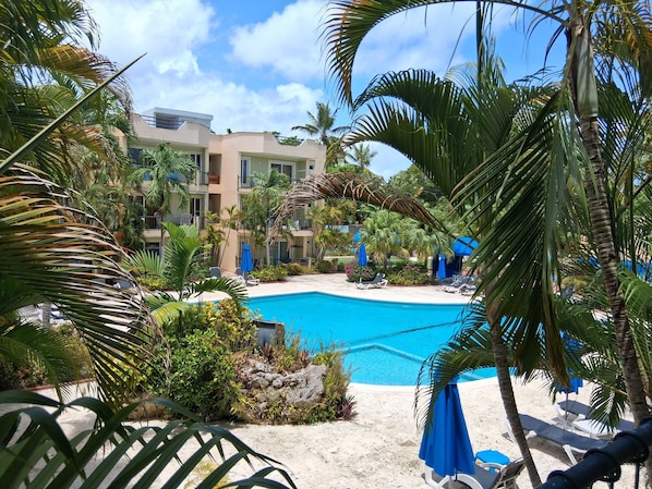 A pool area with lounge chairs and umbrellas, surrounded by palm trees and a building with balconies.