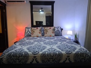 Ein Bett mit einem gemusterten Überwurf, eine Nachttischlampe, ein Topfpflanze und ein Spiegel über dem Bett.