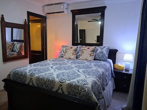 A bedroom with a bed, a nightstand, a lamp, an air conditioning unit, a mirror, and a door.