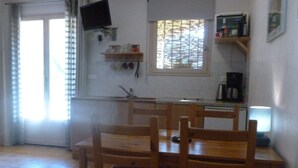 Une cuisine avec des chaises en bois, un téléviseur fixé au mur et une fenêtre avec des stores.