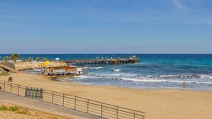 Ein Strand mit einer hölzernen Pier, die sich ins Meer erstreckt, einem Sandweg und einem wolkenlosen, blauen Himmel.