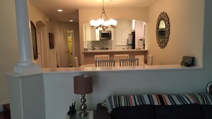 A modern living room with a dark-colored sofa, a glass coffee table, and a lamp. There is a kitchen area with a microwave, stove, and dining table with chairs. A decorative mirror hangs on the wall. - STEPS FROM THE BEACH - HUNTINGTON CONDO - BEAT THE HOTEL COSTS (Huntington Beach)