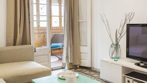 Una sala de estar con sofá, televisor sobre un mueble bajo, una mesita con un plato y una vela, y vista al exterior a través de una puerta.