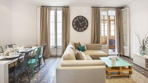 Una sala de estar con un sofá, una mesa de comedor con sillas, un reloj en la pared y una ventana con cortinas.