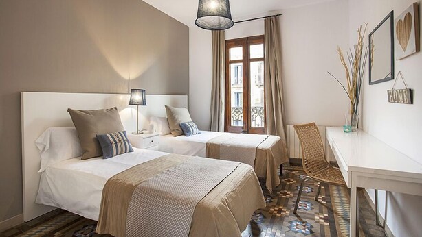 Habitación de hotel con dos camas, un escritorio y una silla. Se observa una ventana con cortinas, una lámpara colgante y adornos en la pared.