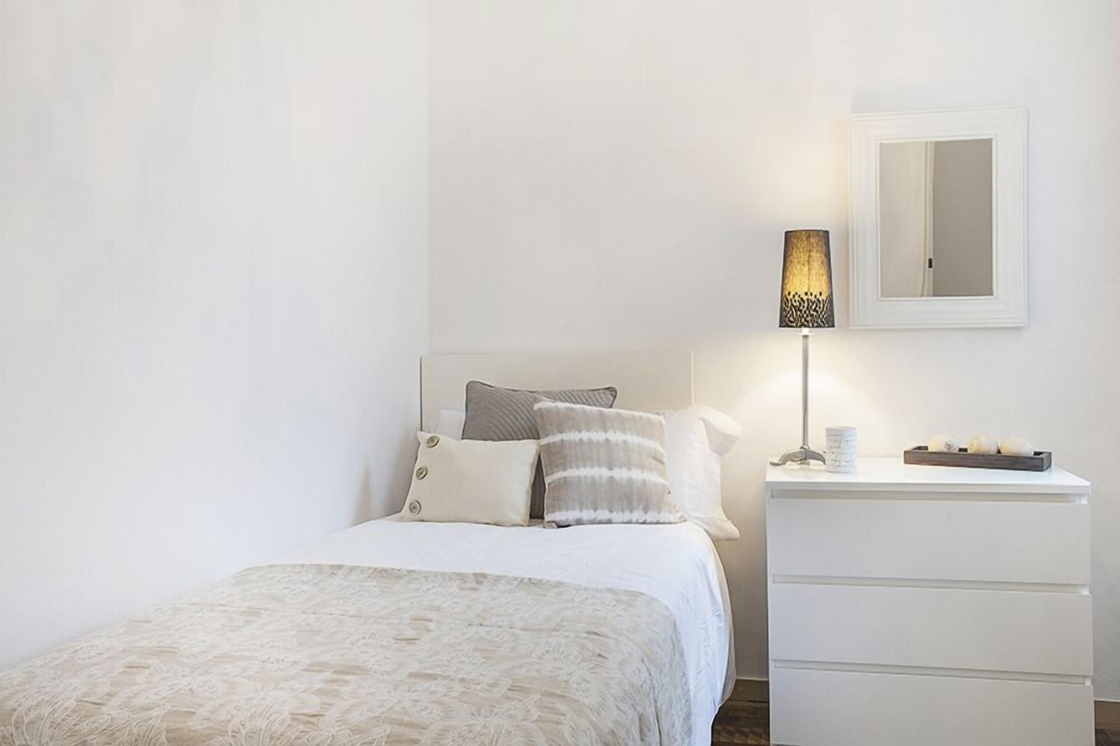 Un dormitorio con cama, almohadas, mesita de noche con lámpara y espejo en la pared.