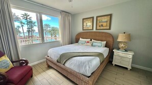 Un dormitorio con un ventanal grande, cama de mimbre, dos cuadros enmarcados, una lámpara de noche y una silla.