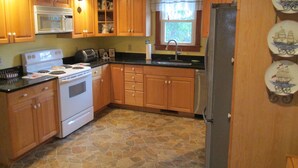 A kitchen with wooden cabinets, a white stove, a refrigerator, and a window with a view of greenery outside. - Cottage with Harbor and Chesapeake Bay View in Rock Hall, MD (Rock Hall)