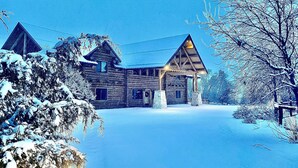 Une cabane en rondins avec un toit triangulaire et une véranda, entourée d’arbres couverts de neige.