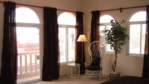 Une chambre d’hôtel avec de grandes fenêtres en arc de cercle, une chaise, une lampe et une plante en pot.