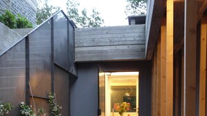 Un estrecho sendero de concreto que lleva a una puerta de madera, delimitado por una cerca de metal y una jardinera con plantas en flor.