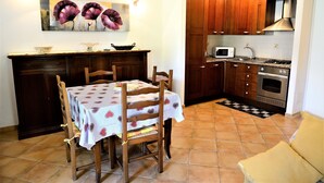 Un comedor con una mesa dispuesta para cuatro personas, sillas de madera, una cocina con armarios de madera y electrodomésticos de acero inoxidable, y un cuadro de flores en la pared.