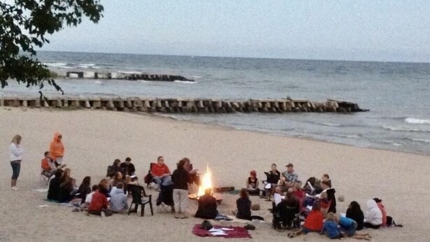A beach with people gathered around a fire, some sitting and some standing, with the ocean and a rocky breakwater in the background. - Last-minute Labor Day plans?  Spend the holiday on the beach in Wisconsin! (Sheboygan)