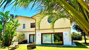 Une maison à deux étages dotée d’une grande porte et de grandes fenêtres en verre, entourée d’une pelouse bien entretenue et de plantes tropicales.