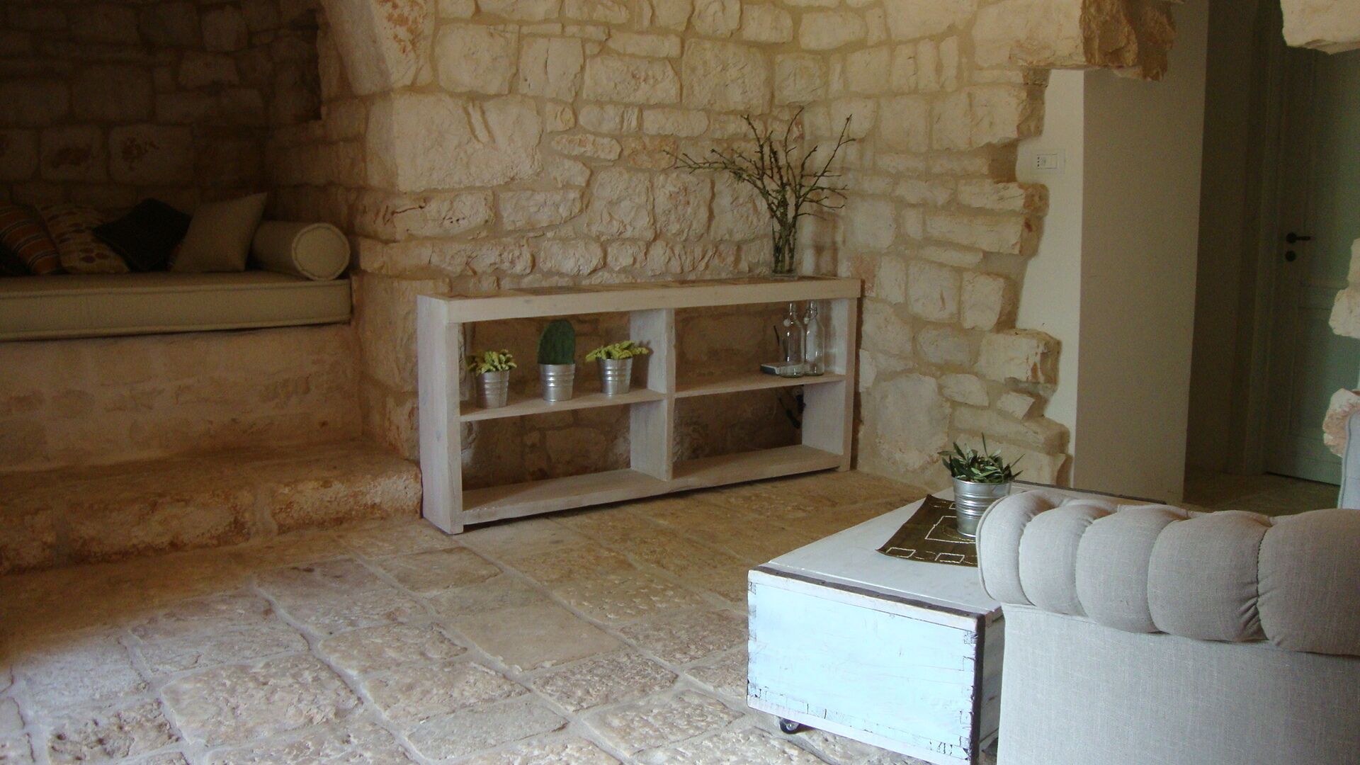 Una habitación con paredes de piedra, una consola blanca con plantas en macetas, un banco acolchado y un armario blanco con ruedas.