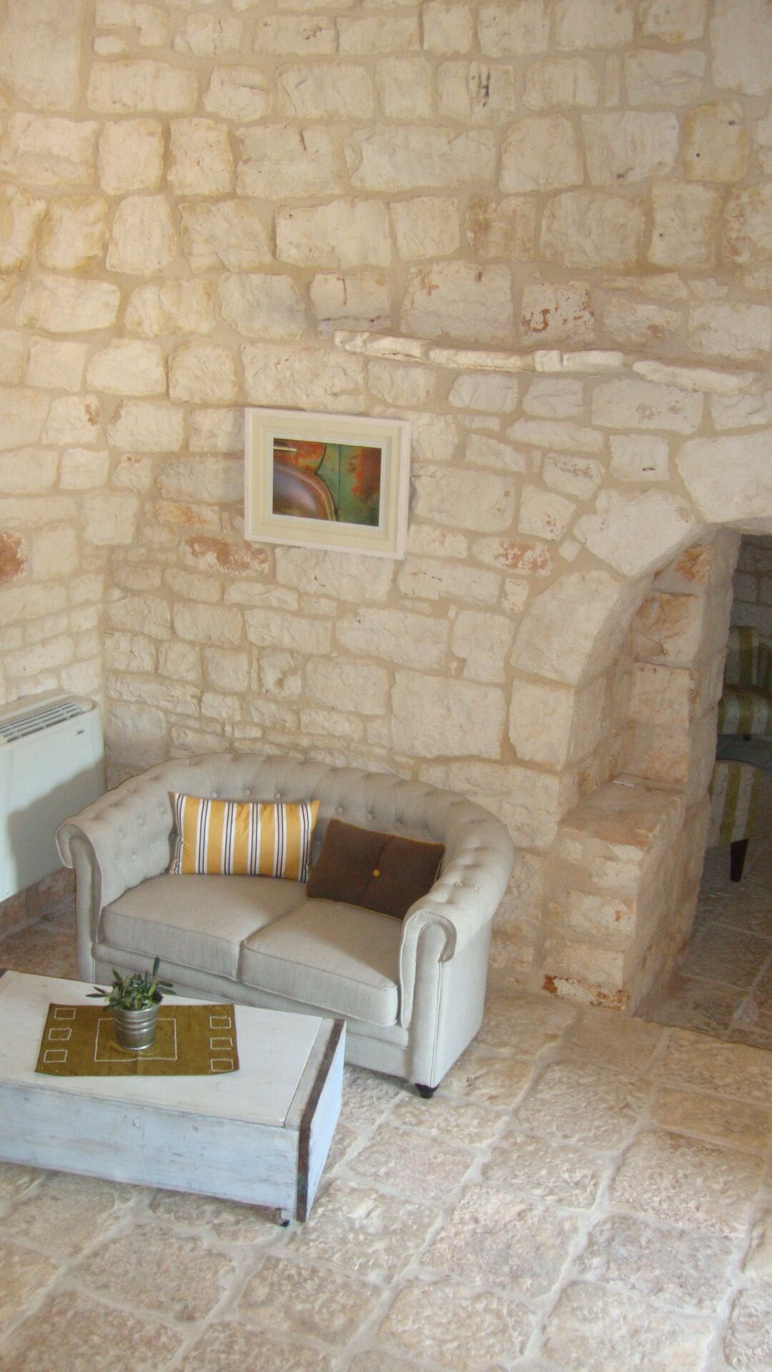A room with a stone wall, a grey sofa with striped pillows, a small table with a plant, and a framed picture on the wall.