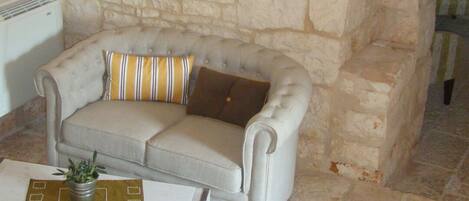 A room with a stone wall, a grey sofa with striped pillows, a small table with a plant, and a framed picture on the wall.