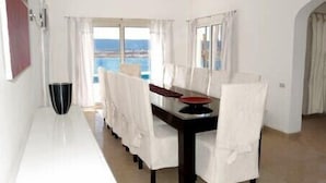 A dining area with a long table, white chairs, and a view of the sea through large windows.