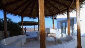 A circular outdoor seating area with a thatched roof, surrounded by stone walls, and a view of a mountainous landscape.