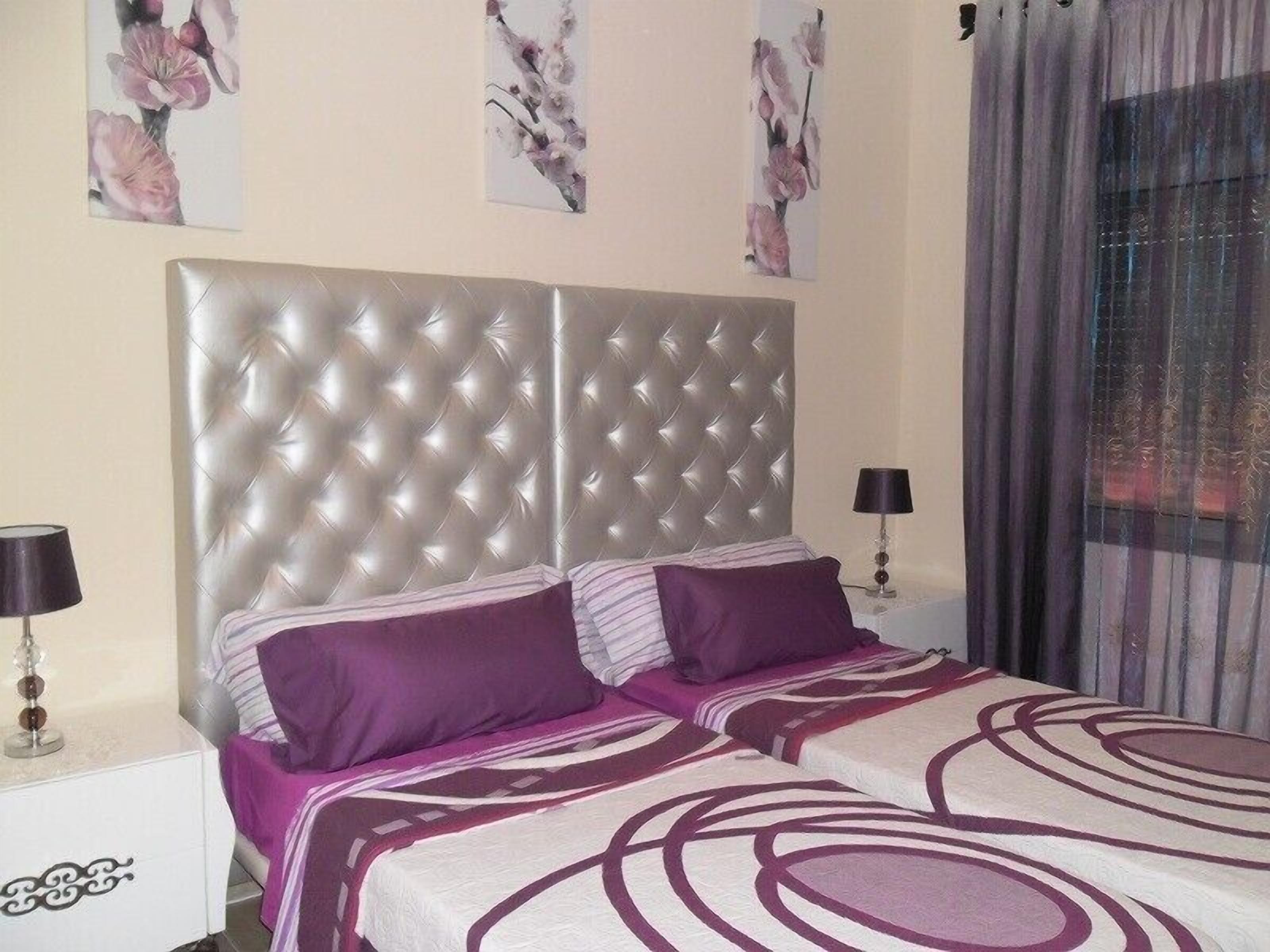 A bedroom with a tufted headboard, two beds with purple bedding, a nightstand with a lamp, and floral artwork on the wall.