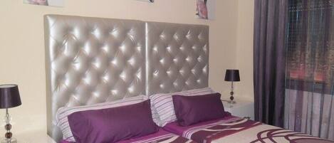 A bedroom with a tufted headboard, two beds with purple bedding, a nightstand with a lamp, and floral artwork on the wall.