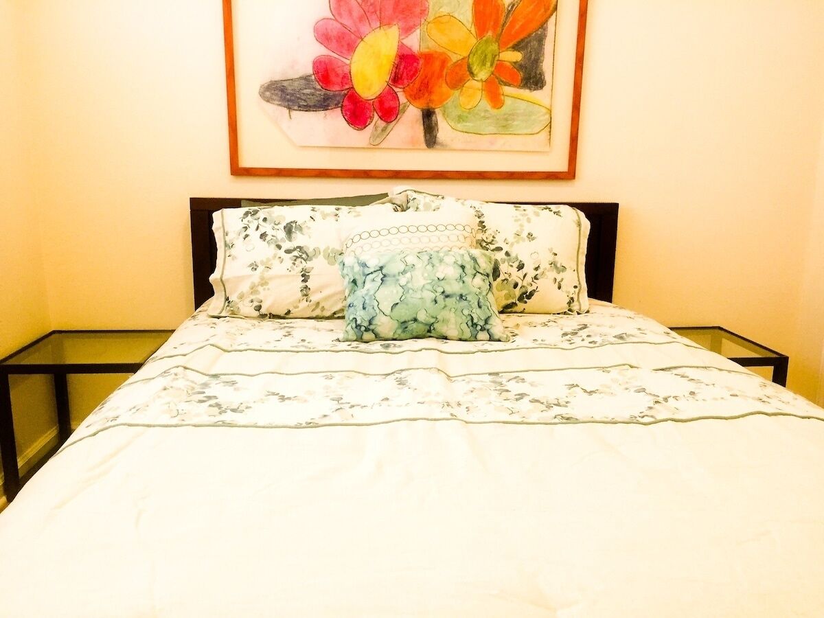 Una cama con una colcha y almohadas estampadas con motivos florales, un cuadro enmarcado de flores en la pared y una mesita de cristal al lado de la cama.