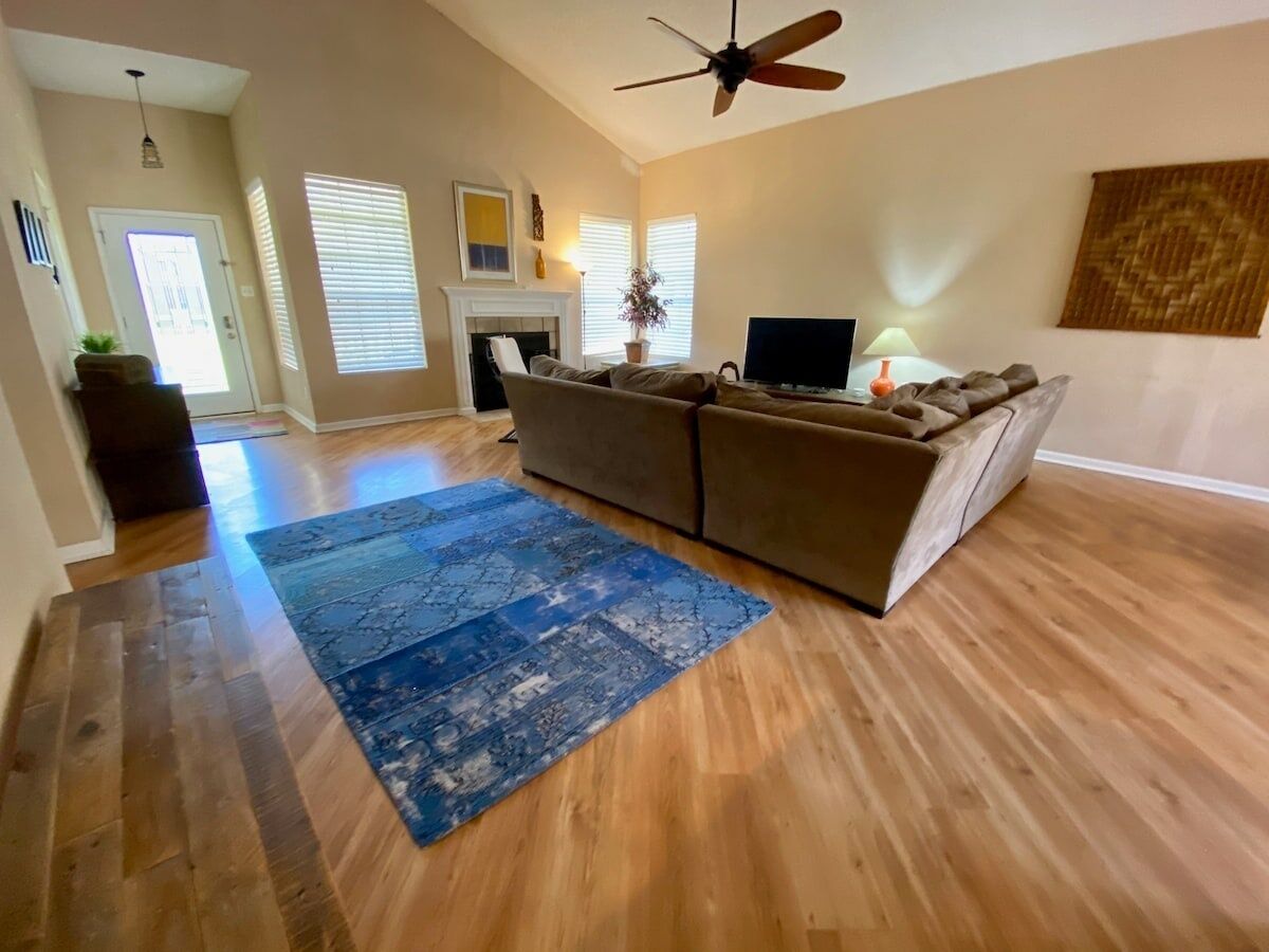 A spacious living room with hardwood flooring, a large sectional sofa, a flat-screen TV, and a ceiling fan.