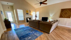 A spacious living room with hardwood flooring, a large sectional sofa, a flat-screen TV, and a ceiling fan.