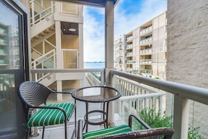 A balcony with two wicker chairs and a small table, overlooking a building with multiple balconies and a view of the water.