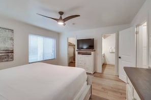 A bedroom with a bed, a ceiling fan, a TV on a stand, and a door leading to another room.