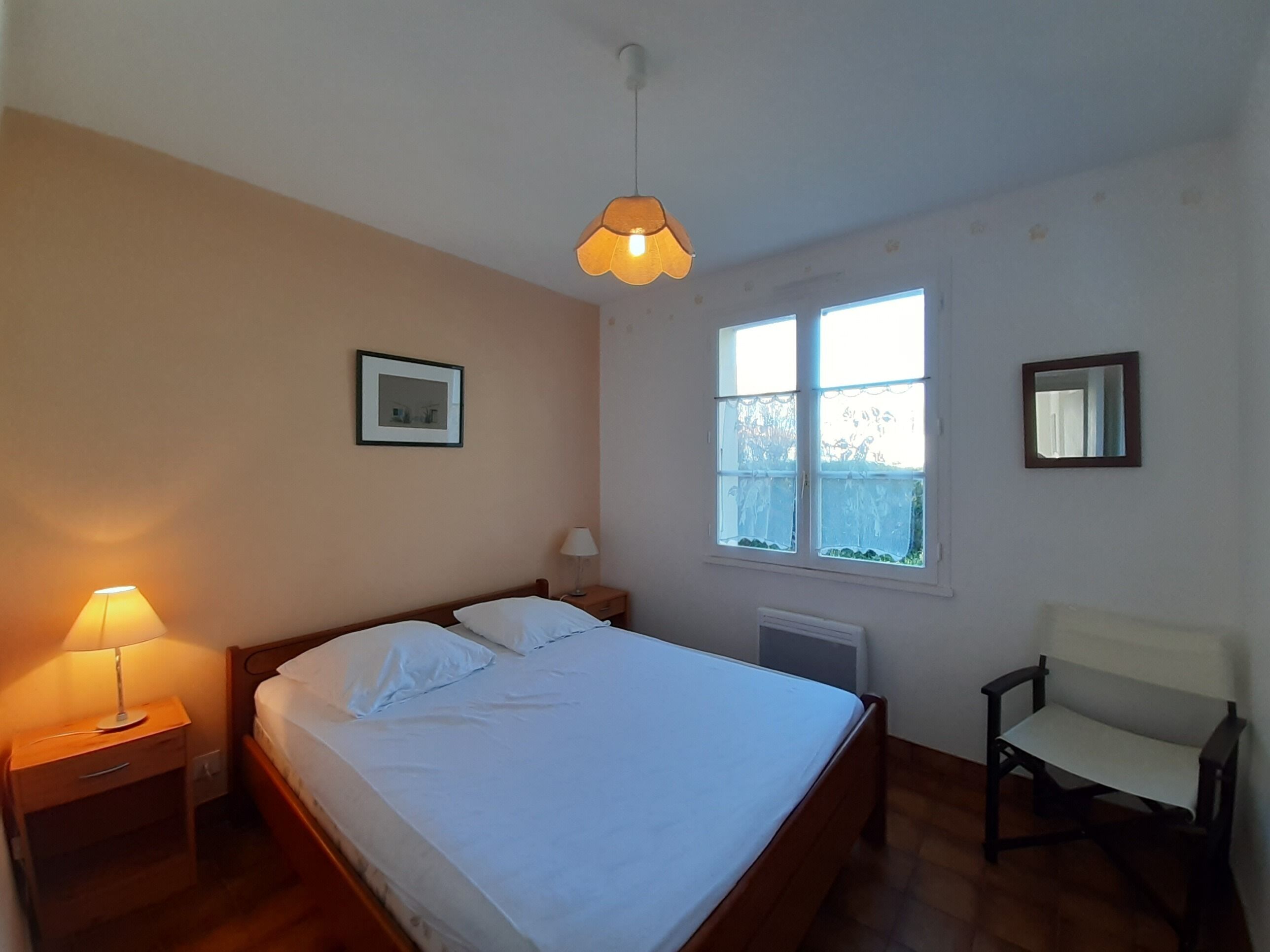 A bedroom with a bed, bedside lamp, chair, window, and a picture on the wall.