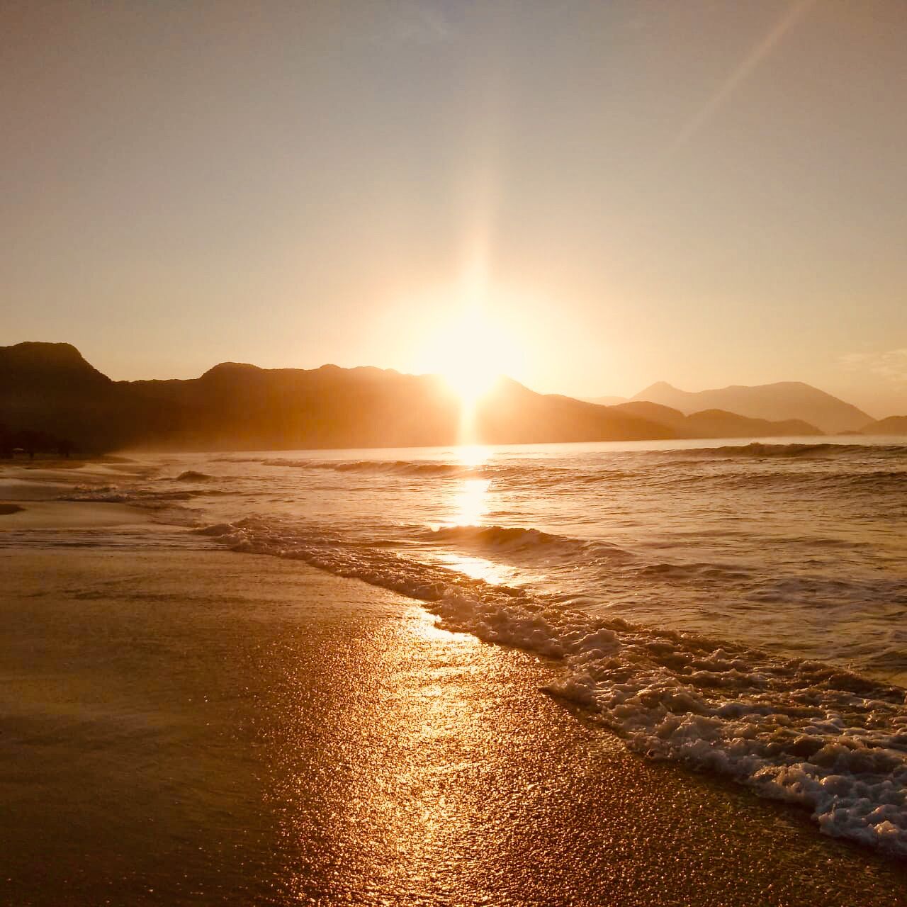 A beach scene with the sun setting over the water, creating a reflective surface on the sand.