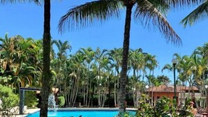 A pool surrounded by palm trees and a building with a tiled roof. - 1 BEDROOM APARTMENT, COND. CLOSED, FACING THE POOLS AND 50M FROM THE BEACH (São Sebastião)