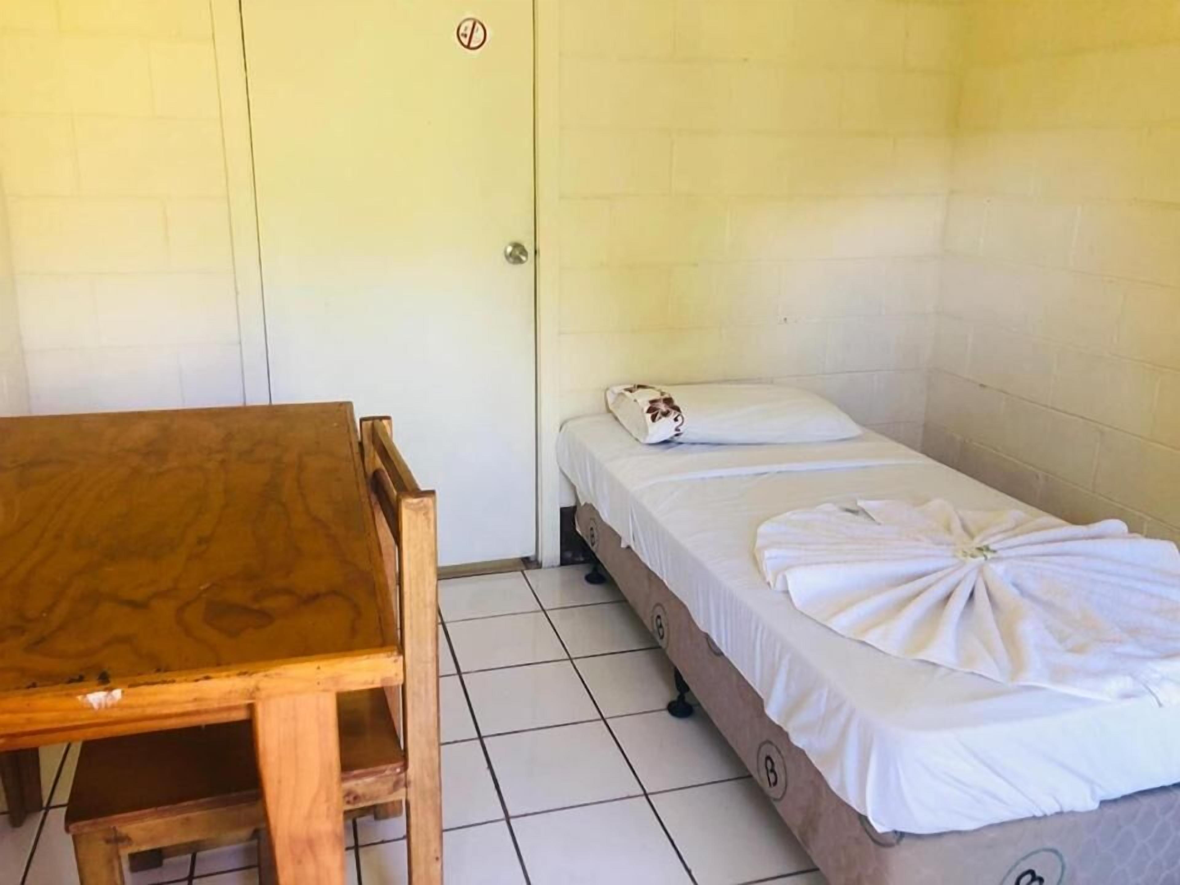Een kleine, schone kamer met een houten tafel en een enkel bed met een net opgemaakt wit laken en een decoratief kussen.