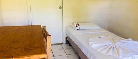 Una habitación pequeña y limpia con una mesa de madera y una cama individual con una colcha blanca bien tendida y una almohada decorativa.
