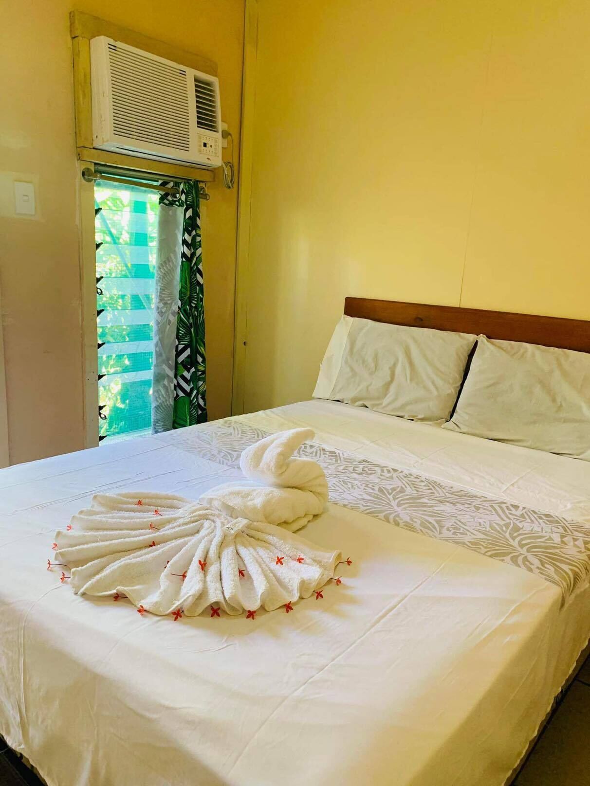 A neatly made bed with a towel swan centerpiece, a window with a view of greenery, and an air conditioning unit mounted on the wall.