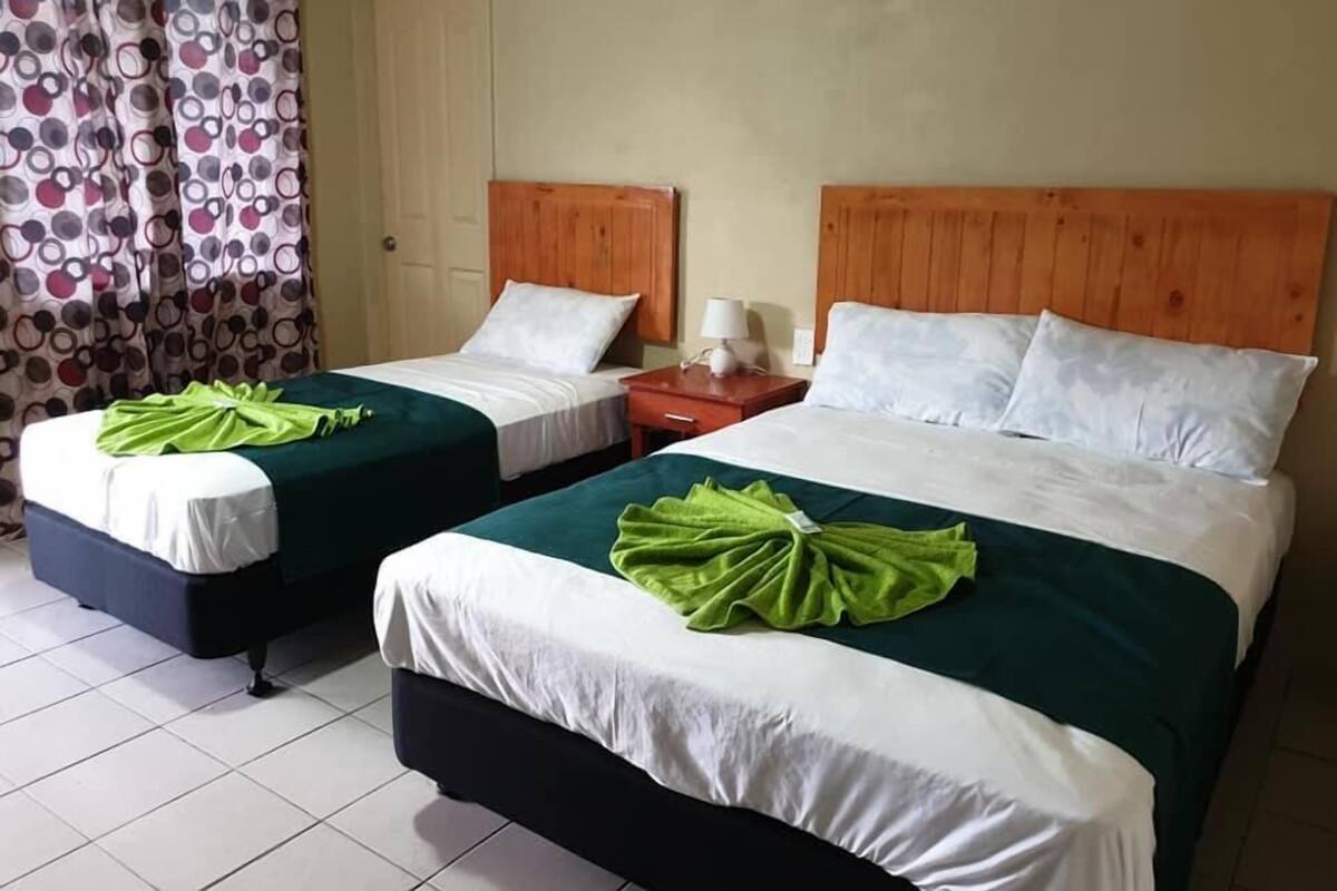 A hotel room with two single beds, each with a green towel and white bedding. There is a wooden headboard, a nightstand with a lamp, and a curtain with a circular pattern.