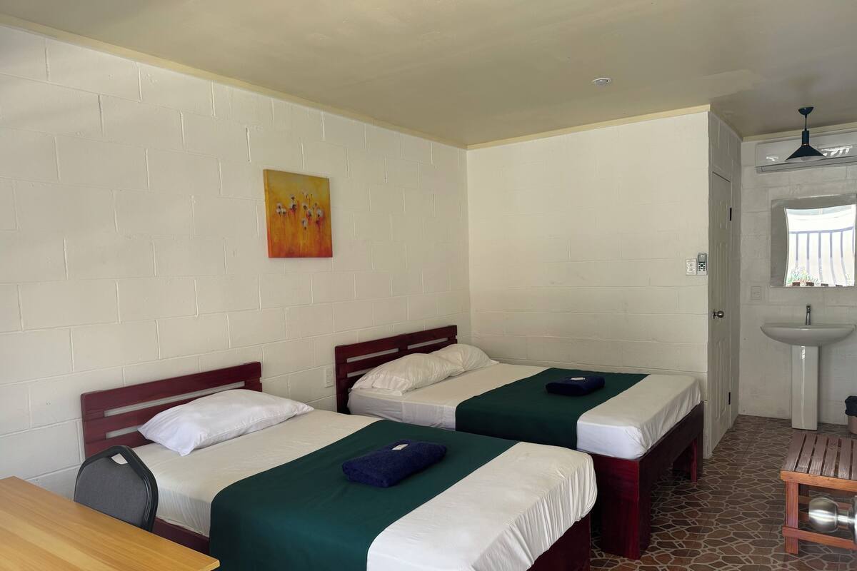 A room with two beds, a wooden table, a bench, a sink, and a painting on the wall.