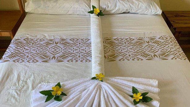 A neatly made bed with a towel arranged in a fan shape and a decorative flower. - Transit Motel - Hostel (Mulifanua)