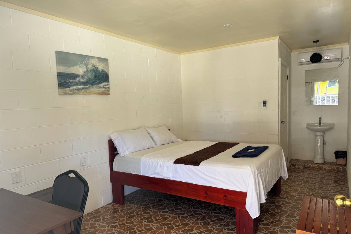 A room with a bed, a desk, a chair, a sink, and a painting on the wall.