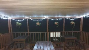 An outdoor wooden deck with picnic tables and benches, string lights, and hanging plants. - Transit Motel - Hostel (Mulifanua)