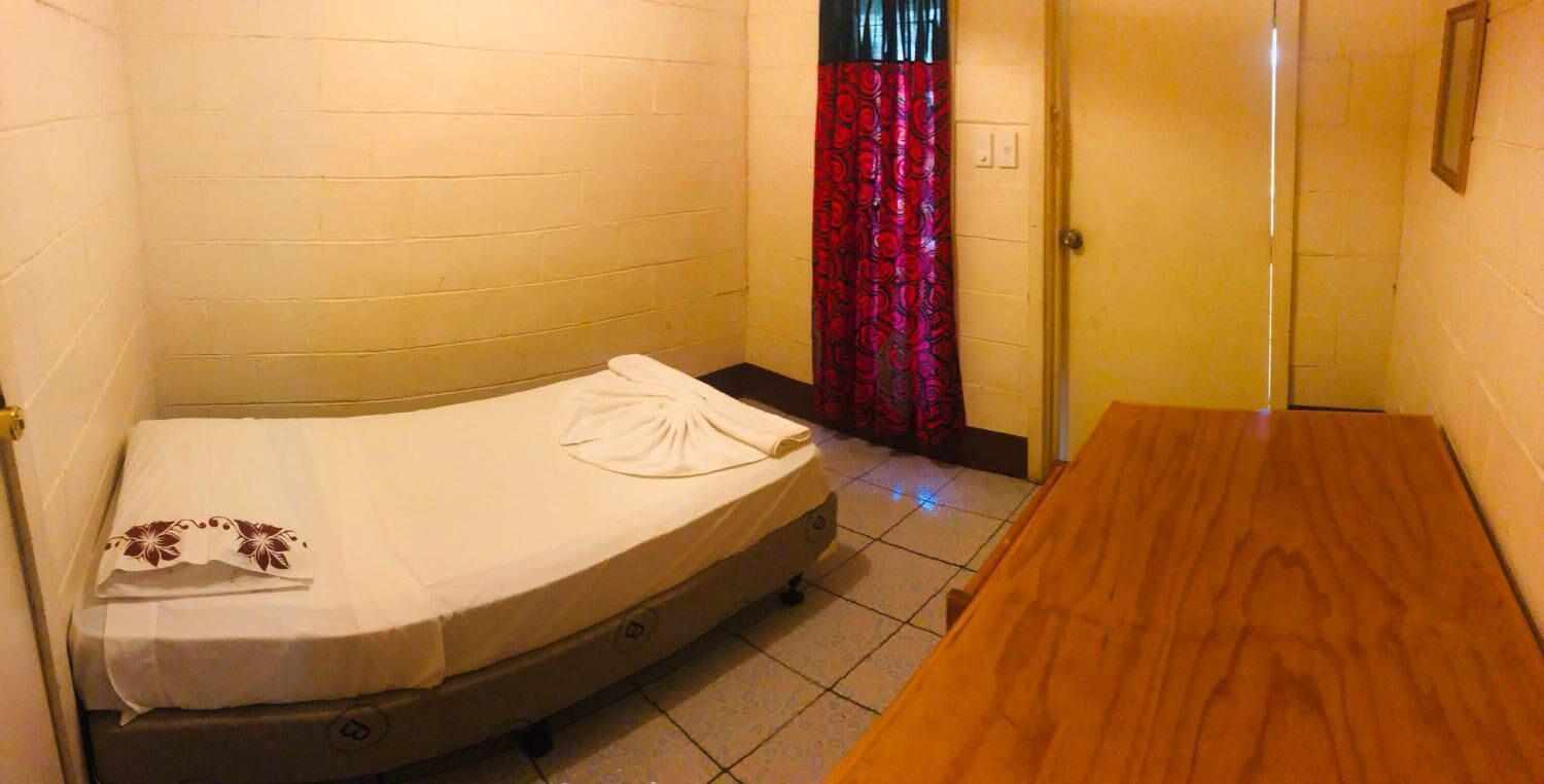 A small, single-bed room with a wooden table, a white mattress, and a red patterned curtain.